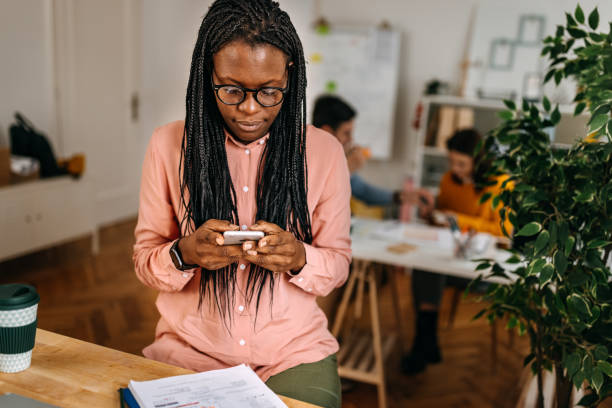 Female design professional with dreadlocks using smart phone while sitting at desk in small office